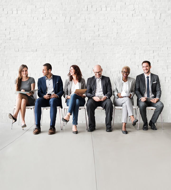 Sechs Personen in Businesskleidung sitzen lächelnd auf Stühlen vor weißer Ziegelwand.